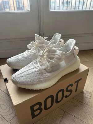 Vita Yeezy Boost 350 V2 sneakers - Snygga vita Yeezy Boost 350 V2 sneakers med stickad ovandel i mesh och klassisk tjock, räfflad sula. Skorna har rund tå, vita skosnören och en modern, sportig look. Perfekta för dig som gillar streetwear och vill ha något stilrent.