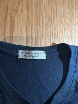 Svart Burberry t-shirt med tryck - Snygg svart t-shirt från Burberry med klassiskt vitt tryck på bröstet där det står 'BURBERRY LONDON ENGLAND PARIS'. T-shirten har rund hals och korta ärmar. Perfekt för dig som gillar stilrena och exklusiva plagg.