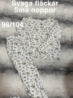 Blommig vit pyjamas med volang - Söt tvådelad pyjamas i vitt med små blommor. Tröjan har långa ärmar och volangdetalj vid axlarna, byxorna har resår i midjan. Perfekt för mysiga kvällar hemma.🌸 Svaga fläckar. Små noppor. 98/104
