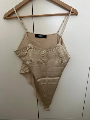 Beige body från Primark med tunna band - Snygg beige body från Primark med tunna axelband och markerade kupor. Tillverkad i mjukt stretchigt material som sitter skönt mot kroppen. Perfekt att styla under en skjorta eller kavaj för en trendig look.