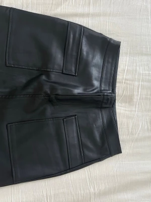 Svart kortkjol i skinnimitation från Amisu - Säljer en svart kortkjol i skinnimitation från Amisu. Kjolen har stora, snygga fickor framtill och en clean, modern look. Perfekt för dig som vill ha en edgy och trendig stil.