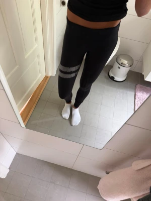Svarta leggings med vita ränder - Säljer ett par svarta leggings med två vita ränder runt ena låret. De har en tight passform och är perfekta för en sportig och avslappnad stil. Materialet ser stretchigt och bekvämt ut, vilket gör dem smidiga att röra sig i.