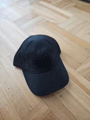 Svart Miu Miu denim keps med broderad text - Svart Miu Miu keps i klassisk modell med böjd skärm och broderad text framtill i samma färg som kepsen. Tillverkad i bomullstyg och har justerbar rem baktill för perfekt passform. Enkel och stilren design som passar till många outfits.
