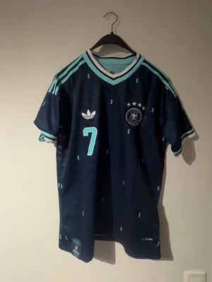 Mörkblå fotbollströja Tyskland #7 - Snygg mörkblå fotbollströja med ljusblå detaljer och nummer 7, samt namnet Havertz på ryggen. Tröjan har korta ärmar, V-ringning och klassiska Adidas-ränder på axlarna. Perfekt för fotbollsträning eller match.