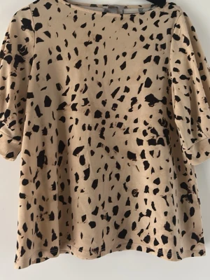 Beige topp med svart leopardmönster - Trendig beige topp med svart leopardmönster och halvlånga puffärmar. Toppen har en avslappnad passform och rund halsringning. Perfekt för dig som vill sticka ut med ett coolt mönster och en modern siluett.