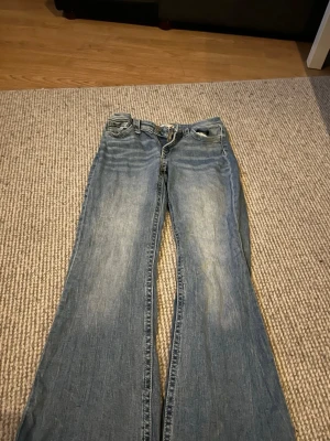 Blå bootcut jeans med slitningar - Snygga blå bootcut jeans med lätt slitna detaljer framtill och bakfickor med dekorativa sömmar. Jeansen har klassisk femficksdesign och är tillverkade i denim. Perfekta för en avslappnad och trendig look. Den har lite defekter som syns på bilderna (slitna i benen vid fötterna och stenar saknas) pris diskuteras 