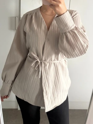 Beige plisserad blus med knytband - Stilren beige blus med plisserat tyg och V-ringning. Blusen har långa ärmar med breda muddar och ett smalt knytband i midjan för en snygg siluett. Perfekt att matcha med både jeans och kjol för en trendig look.