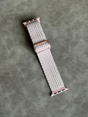 Rosa klockarmband i textil - Snyggt ljusbeige klockarmband i flätad textil med rosa detaljer vid fästena. Justerbart spänne i metall för perfekt passform. Passar till smartklockor och ger en stilren och modern look.