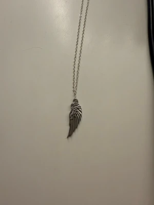 Halsband med silverfärgad vinge - Snyggt halsband med en detaljrik vinge som hänge i silverfärgad metall. Kedjan är tunn och stilren, perfekt för dig som gillar smycken med en unik touch och symbolik. Passar till många olika stilar och är lätt att matcha med andra accessoarer. Smycket är egengjort i rostfritt stål.