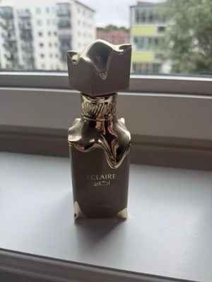 Eclaire lyxig parfymflaska guld - Snygg parfymflaska med namnet Eclaire i elegant design. Flaskan är mörk med detaljer i glänsande guld och har en unik, konstnärlig form på korken. Perfekt för dig som gillar exklusiva och stilrena dofter.