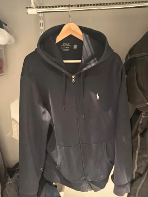 Mörkblå hoodie från Polo Ralph Lauren - Snygg mörkblå hoodie med dragkedja från Polo Ralph Lauren. Klassisk design med huva och dragsnören, två fickor framtill och den ikoniska vita logon broderad på bröstet. Tillverkad i mjukt bomullsmaterial som är skönt att bära.