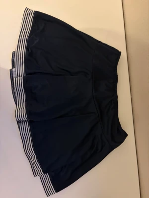 Mörkblå skort från FILA - Sportig mörkblå tenniskjol från FILA med vita och blå ränder längst ner. Kjolen är gjord i ett stretchigt funktionsmaterial som passar perfekt för träning eller tennis. Klassisk design med elastisk midja och snygg passform.DM FÖR FLER BILDER 💕