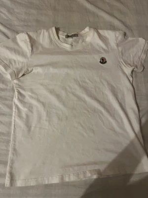 Vit Moncler T-shirt  - Klassisk vit t-shirt från Moncler med rund hals och korta ärmar. På bröstet sitter Monclers ikoniska logga som en snygg detalj. Perfekt basplagg i mjuk bomull som funkar till allt.