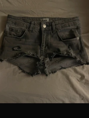 Svarta slitna jeansshorts - Säljer ett par svarta jeansshorts med råa, fransiga kanter och slitningar framtill. Shortsen har fem fickor, knappgylf och en tight passform. Perfekta för dig som gillar en edgy och avslappnad stil. 