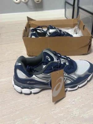 Asics blå och vita sneakers - Snygga sneakers från Asics i blått, vitt och silver. Skorna har mesh- och syntetmaterial för bra andningsförmåga och komfort. Klassisk låg modell med tydliga Asics-loggor på sidan och tungan. Perfekta för dig som gillar sportig stil.