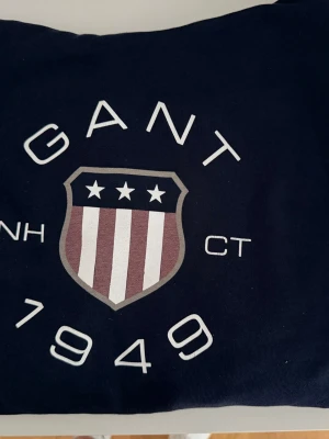 Mörkblå hoodie från Gant - Snygg mörkblå hoodie från Gant med stort tryck framtill som visar Gant-loggan och texten '1949'. Hoodien har huva med dragsko och en stor magficka. Perfekt för en avslappnad och sportig stil.