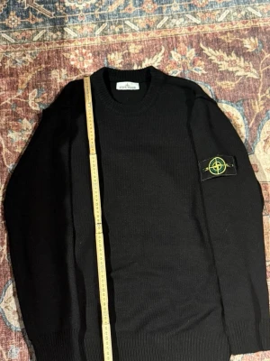 Svart stickad tröja från Stone Island - Snygg svart stickad tröja från Stone Island med klassisk rund halsringning och ribbade muddar. Tröjan har den ikoniska Stone Island-loggan på vänster ärm. Perfekt för dig som gillar stilrena och tidlösa plagg.