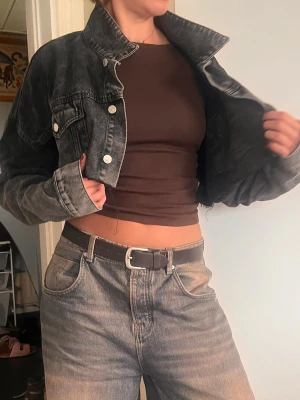 Mörkgrå croppad jeansjacka - Trendig croppad jeansjacka i svart tvättad denim med silverfärgade knappar och två bröstfickor med lock. Jackan har en avslappnad passform och rå kant nedtill för en edgy look. Perfekt att slänga över en t-shirt eller crop top.