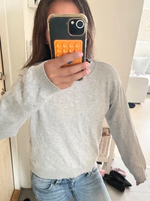 Ljusgrå Tröja - En enkel och stilren ljusgrå tröja med rund hals och långa ärmar. Tröjan har ribbade muddar vid ärmslut och nederkant, perfekt för en clean och avslappnad look. Passar till det mesta och är lätt att matcha med jeans eller kjol.