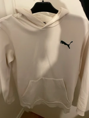 Vit hoodie från Puma med tryck - Snygg vit hoodie från Puma med svart logga på bröstet och stort grafiskt tryck med PUMA-text på ryggen. Klassisk känguruficka framtill och huva. Perfekt för en chill och sportig stil.