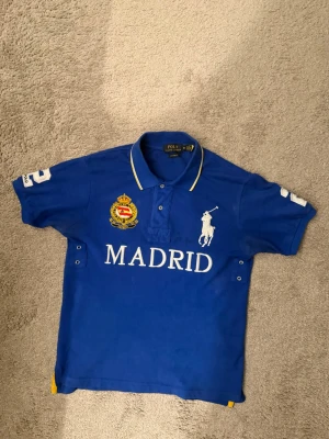 Polo Ralph Lauren Big Pony Madrid Polo Blue - Blå pikétröja med vit text 'MADRID' och stor logga framtill. Har broderad detalj på bröstet, nummer 2 på ryggen och ärmarna samt gul kant på kragen. Klassisk polokrage och korta ärmar. Perfekt för en sportig look.