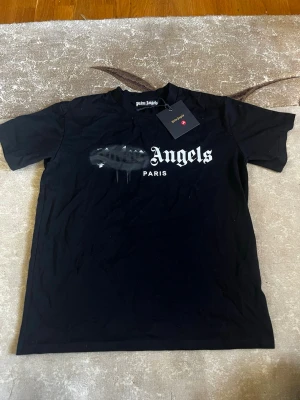 Svart Palm Angels t-shirt Paris - Svart t-shirt från Palm Angels med trycket 'Palm Angels Paris' på bröstet. T-shirten har korta ärmar och en klassisk rund hals. Trycket har en cool spray paint-effekt över 'Palm'. Perfekt för dig som gillar streetwear och vill sticka ut.