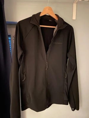 Svart vindjacka med dragkedja   - Svart vindjacka/träningsjacka med hel dragkedja framtill och två fickor med dragkedja. Jackan har en sportig look och är tillverkad i ett lätt syntetmaterial. Perfekt för blåsiga dagar och träning. Hör gärna av er om ni har några frågor eller funderingar! Priset är inte hugget i sten vid snabb affär. 