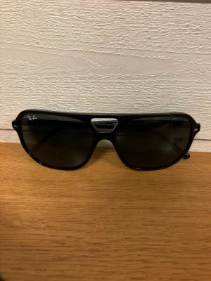 Svarta Ray-Ban pilotbågar solglasögon - Snygga svarta solglasögon från Ray-Ban med klassisk pilotbåge och mörka glas. Kommer med äkthetskort, putsduk och orginalfodral. Perfekta för dig som vill ha en tidlös och trendig look.