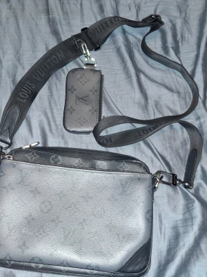 axelväska - Säljer en svart axelväska  med klassiskt monogrammönster och justerbar axelrem. Väskan har en tillhörande liten väska som kan fästas på remmen. Perfekt för dig som vill ha en stilren och praktisk accessoar.
