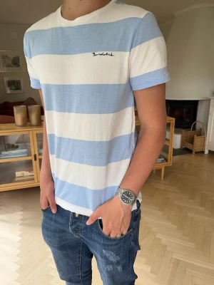 Randig blåvit t-shirt Bondelid - Bondelid T-shirt i klassiska blåvita ränder. Stilren och tidlös – perfekt till sommaren, jeans, shorts eller lager på lager. Mjuk och skön passform med broderad Bondelid-logga på bröstet. Ett måste för dig som gillar preppy, old money och Scandinavian style.  #Bondelid #Tshirt #Randig #Blå #Vit #Preppy #OldMoney #QuietLuxury #ScandinavianStyle #Casual #Streetwear #Basic #Minimalist #Sommar #Herr #Designer #Kvalitet #Mode #Jeans #Trend