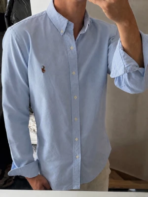 Ljusblå skjorta från Ralph Lauren - Klassisk ljusblå skjorta från Ralph Lauren med button-down krage och broderad logga på bröstet. Skjortan har långa ärmar, vita knappar och är tillverkad i mjuk bomull. Perfekt till jeans eller chinos för en clean look.