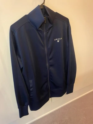 Mörkblå zip hoodie från SikSilk - Mörkblå hoodie från SikSilk med dragkedja framtill och hög krage. Tillverkad i ett glansigt, mjukt material och har två fickor på sidorna. Diskret logga i vitt på bröstet. Perfekt för en chill och sportig look.