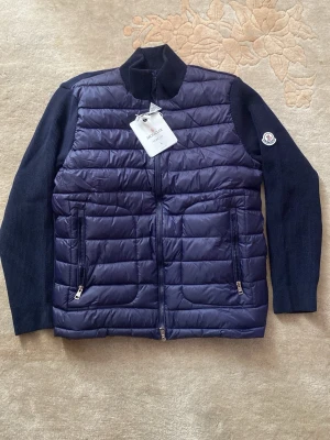 Mörkblå cardiganjacka från Moncler - Snygg mörkblå cardiganjacka från Moncler med quiltad front i glansigt material och stickade ärmar och rygg. Jackan har två dragkedjeförsedda fickor fram, hög krage och Moncler-logga på ärmen. Perfekt för lager-på-lager och stilren look.‼️⚠️⚠️Kvitto finns!!!! ⚠️⚠️⚠️‼️