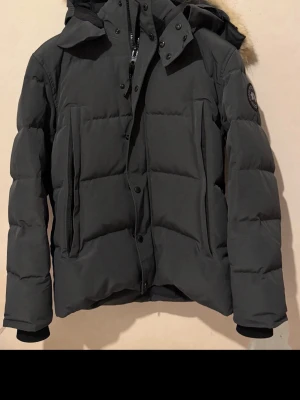 Svart dunjacka med huva och fuskpäls - Snygg svart dunjacka med huva och fuskpälsdetalj. Jackan har en quiltad design, dragkedja och knappar framtill samt två stora fickor. Muddar vid ärmslut för extra värme och en patch på ärmen för en cool touch.