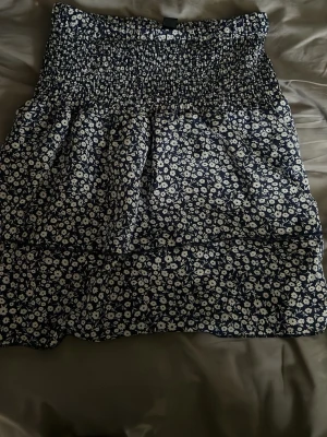 Blommig kortkjol med volang - Supersöt kortkjol med mörkblå botten och vita små blommor. Kjolen har smock i midjan för skön passform och volangdetalj nedtill som ger en extra touch. Perfekt för dig som gillar en feminin och avslappnad stil.