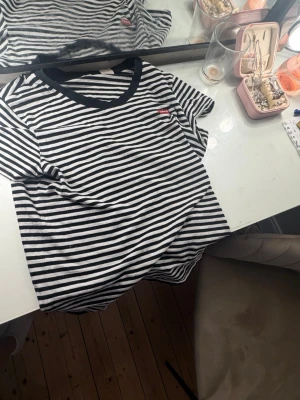 Randig svartvit t-shirt med v-ringning - Säljer en svartvit randig t-shirt med v-ringning och korta ärmar. T-shirten har en liten röd Levis märke vid bröstet och är gjord i mjuk bomull. Perfekt för en chill och stilren look.