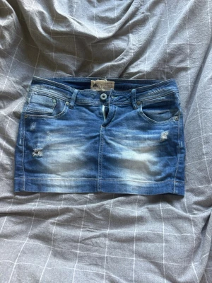 Blå jeanskjol med slitningar - Säljer en blå jeanskjol i kort modell med slitna detaljer och klassiska fickor både fram och bak. Kjolen har en knapp och dragkedja framtill samt ljusa tvättade partier för en cool look. Perfekt för dig som gillar denim och vill ha en trendig stil.