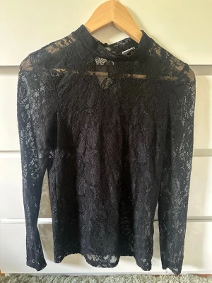 Svart spetsblus med lång ärm - Säljer en svart blus i spets med lång ärm och hög hals. Blusen är genombruten och har ett elegant, feminint uttryck. Perfekt att styla med jeans eller kjol för en trendig look.