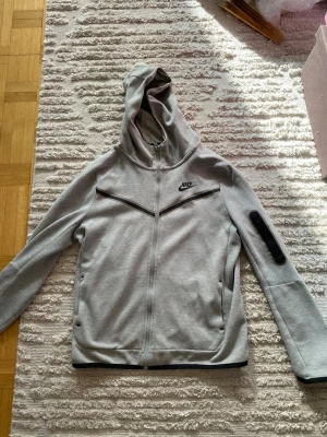 Grå Nike hoodie med dragkedja - Snygg grå hoodie från Nike med hel dragkedja och svart logga på bröstet. Tröjan har huva, svarta detaljer och fickor med dragkedja samt en cool detalj på ena ärmen. Tillverkad i mjukt material, perfekt för chill eller träning.