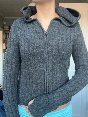 Grå ziphoodie i stickat material - Mysig grå ziphoodie i stickat material med långa ärmar och huva. Tröjan har en smal passform och ribbad struktur som ger en snygg look. Perfekt för lager på lager och chill dagar.