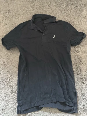 Svart pikétröja från Peak Performance - Klassisk svart pikétröja från Peak Performance med liten vit logga på bröstet. Tröjan har korta ärmar, krage och knappar framtill. Perfekt till jeans eller shorts för en avslappnad stil.