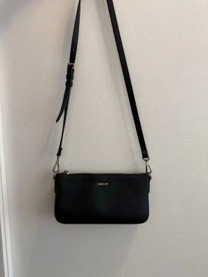 Svart axelväska från DKNY - Snygg svart axelväska från DKNY i slät läderimitation. Väskan har justerbar axelrem, guldfärgade detaljer och dragkedja både fram och bak. Perfekt storlek för det viktigaste och stilren design med DKNY-logga framtill.