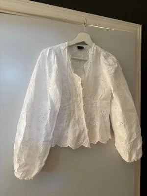 Vit broderad blus med puffärm från Gina - Söt vit blus med broderade blommor och vågig kant. Blusen har puffiga långärmar med resår vid ärmslut och knäppning framtill. Perfekt för en romantisk och trendig look. Materialet känns luftigt och lätt. Strl 36 använd en gång 