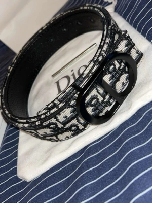 Svartvitt Dior bälte med monogram - Säljer ett svartvitt bälte från Dior med ikoniskt monogrammönster och stor, rundad metallspänne i svart. Bältet har en exklusiv känsla och passar perfekt till att lyfta vilken outfit som helst.