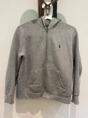 Grå zip-hoodie från Polo Ralph Lauren - Snygg grå hoodie med dragkedja från Polo Ralph Lauren. Klassisk design med två fickor fram, broderad logga på bröstet och huva. Perfekt för en avslappnad och stilren look. Mjuk bomullsblandning och ribbade muddar.