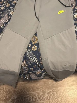 Gråa mjukisbyxor från Nike - Säljer ett par gråa mjukisbyxor från Nike med gul logga på vänster ben. Byxorna har dragsko i midjan och paneldetaljer på benen. Tillverkade i mjukt material för maximal komfort och avslappnad passform.
