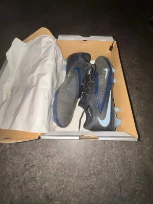 Svarta Nike fotbollsskor med blå detaljer - Säljer ett par snygga svarta Nike fotbollsskor med ljusblå detaljer och dobbar. Skorna har en modern design med stickad ovandel och snörning för bra passform. Perfekta för dig som vill ha stilrena och funktionella skor på planen.