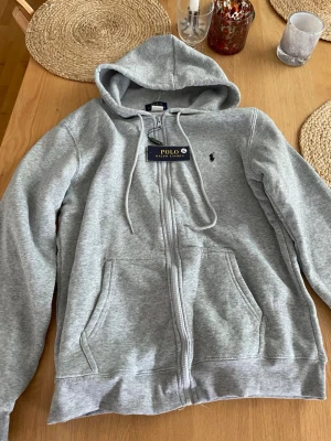 Polo Ralph Lauren hoodie  - Snygg grå hoodie med dragkedja från Polo Ralph Lauren. Klassisk design med huva, dragsnören och två fickor framtill. Liten broderad logga på bröstet. Perfekt för en chill och stilren look. Riktigt sköna. Storlek M. Kom privat vid minsta funderingar!