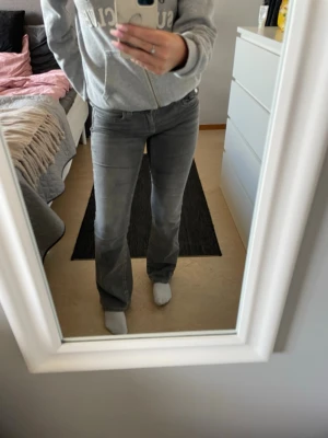 Grå bootcut jeans med fickdetaljer - Säljer ett par gråa bootcut jeans med klassisk femficksmodell och knappdetalj på bakfickan. Jeansen har normal midja och är tillverkade i ett stretchigt denimtyg som sitter snyggt på benen. Perfekta för en avslappnad och trendig look.