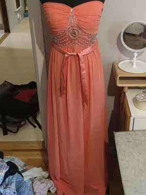 Strapless korallfärgad maxiklänning - Snygg axelbandslös maxiklänning i korall med transparent midjeparti dekorerat med glittriga stenar och pärlor. Klänningen har ett band i midjan som knyts till rosett och ett lätt, flytande tyg som ger en elegant look. Storlek 10 är en brittisk storlek (UK 10)